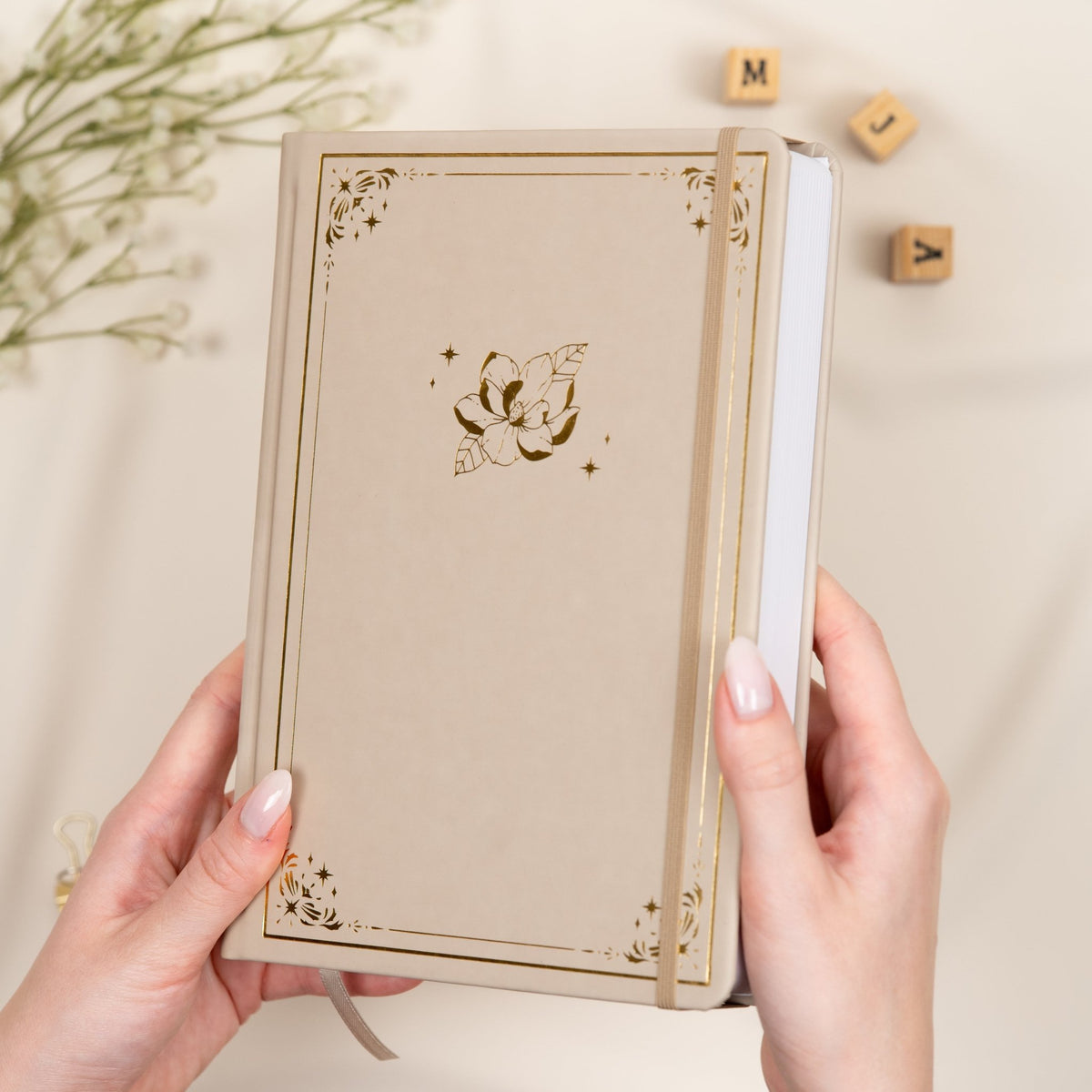 Holding a beige Oat premade dotted bullet style journal made of Italian vegan leather with an embossed gold magnolia design.