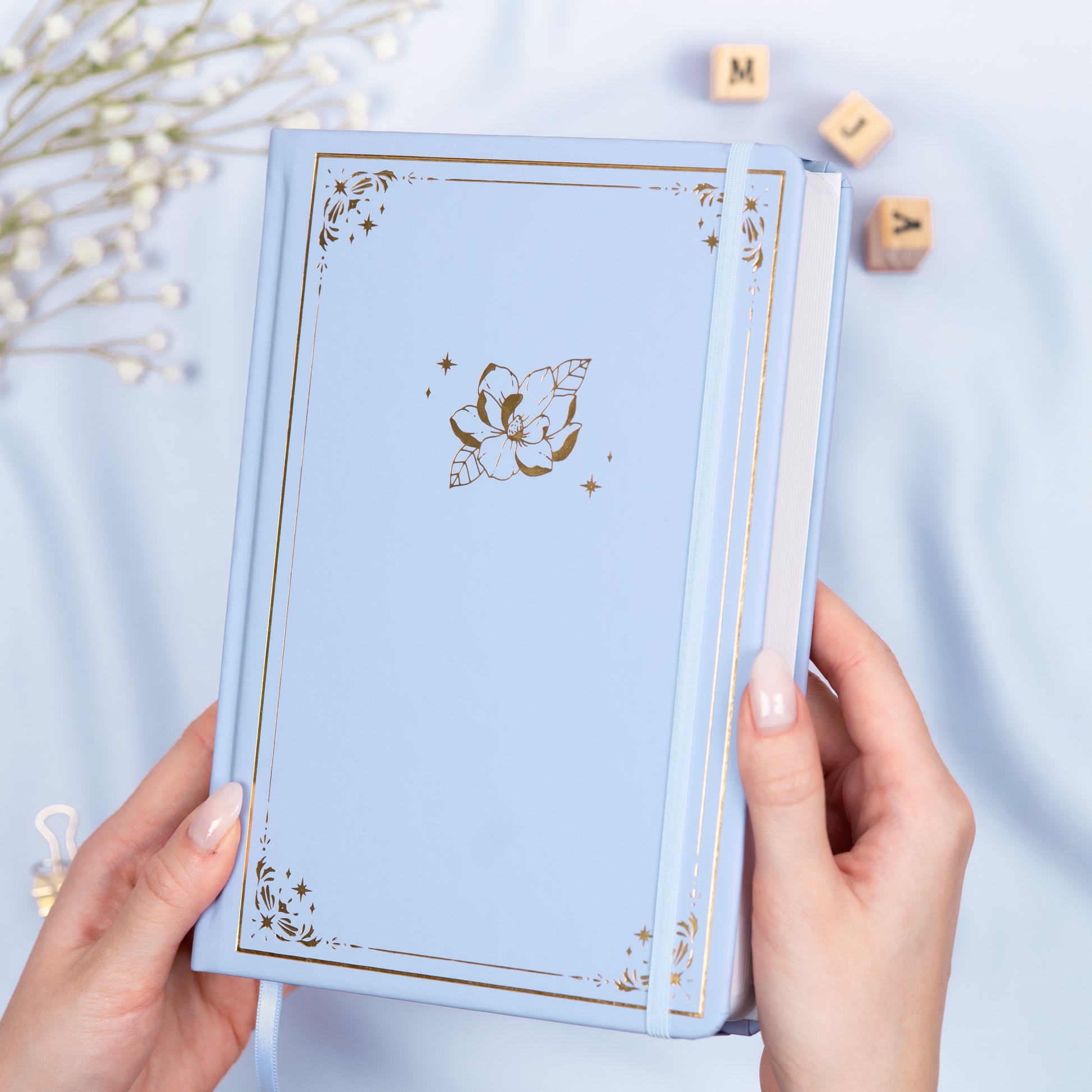 Holding a light blue premade dotted bullet style journal made of Italian vegan leather with an embossed gold magnolia design.