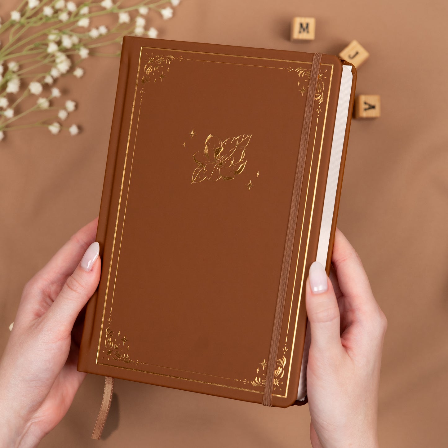 Holding a cinnamon brown premade dotted bullet style journal made of Italian vegan leather with an embossed gold magnolia design.