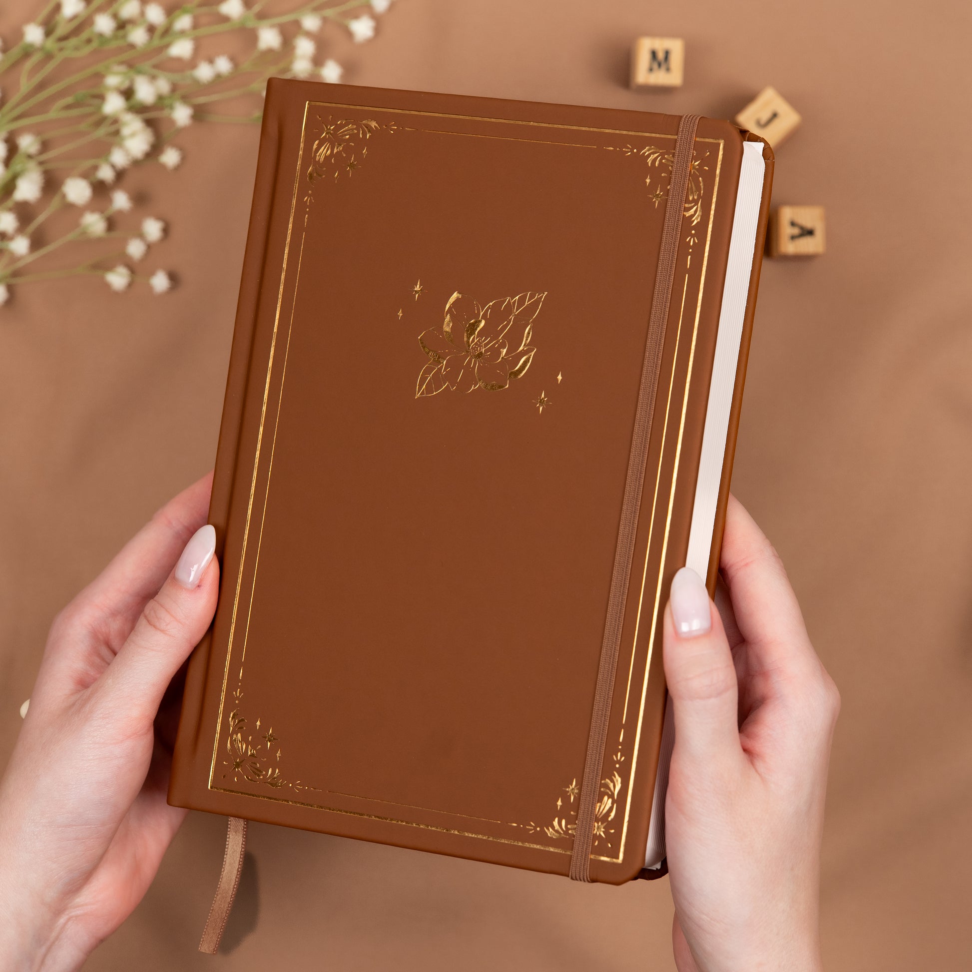 Holding a cinnamon brown premade dotted bullet style journal made of Italian vegan leather with an embossed gold magnolia design.