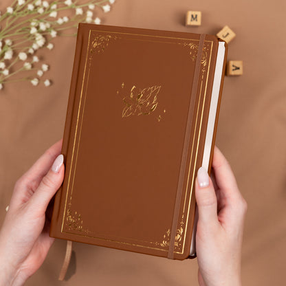 Holding a cinnamon brown premade dotted bullet style journal made of Italian vegan leather with an embossed gold magnolia design.