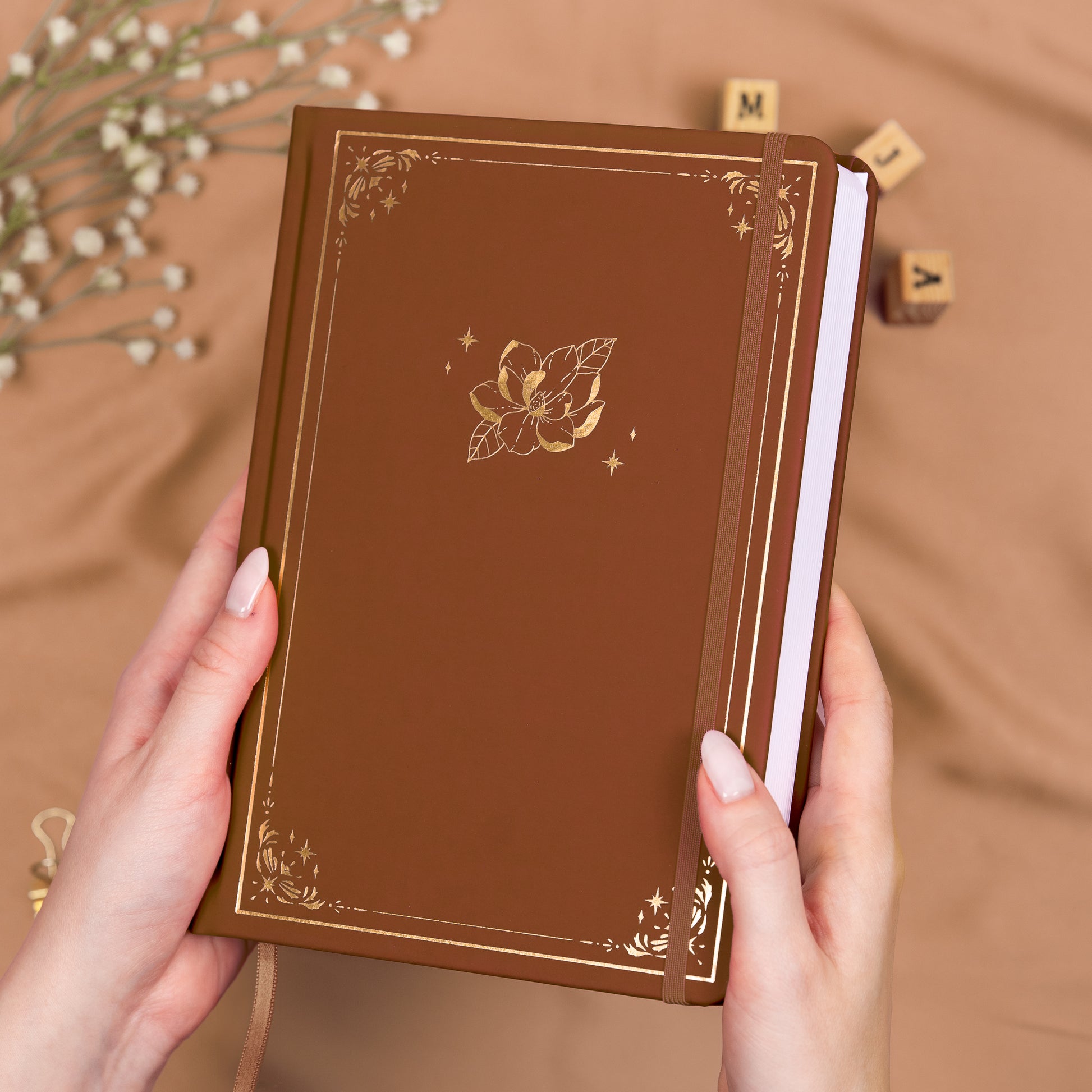 Holding a cinnamon brown premade dotted bullet style journal made of Italian vegan leather with an embossed gold magnolia design.