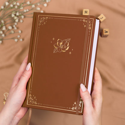 Holding a cinnamon brown premade dotted bullet style journal made of Italian vegan leather with an embossed gold magnolia design.