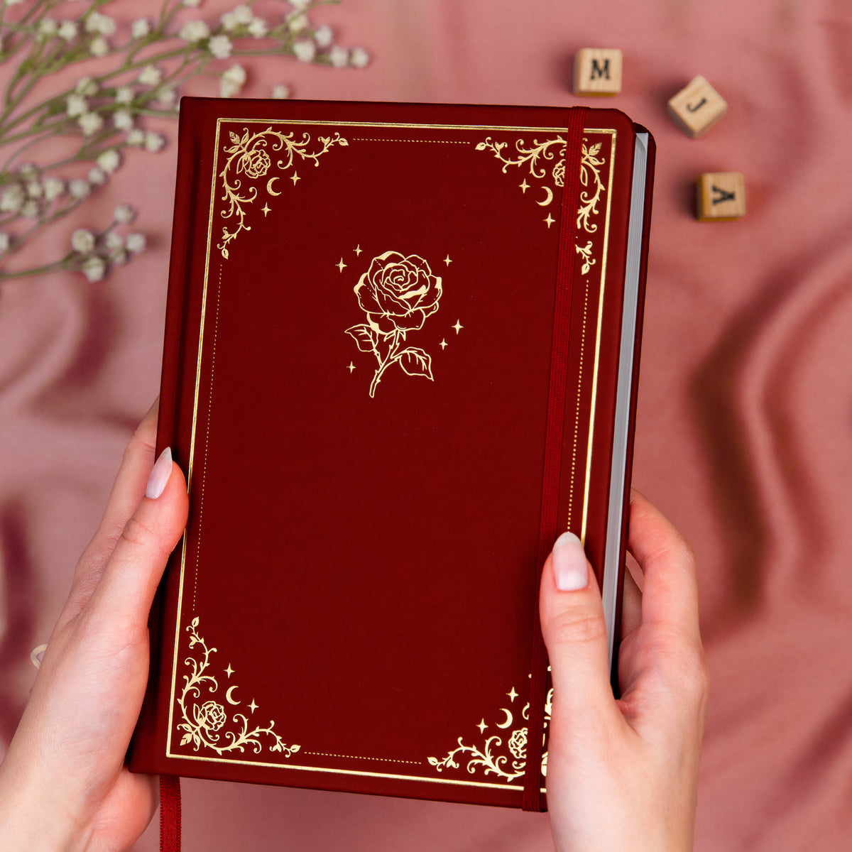 Holding a deep red premade dotted bullet style journal made of Italian vegan leather with an embossed gold rose design.